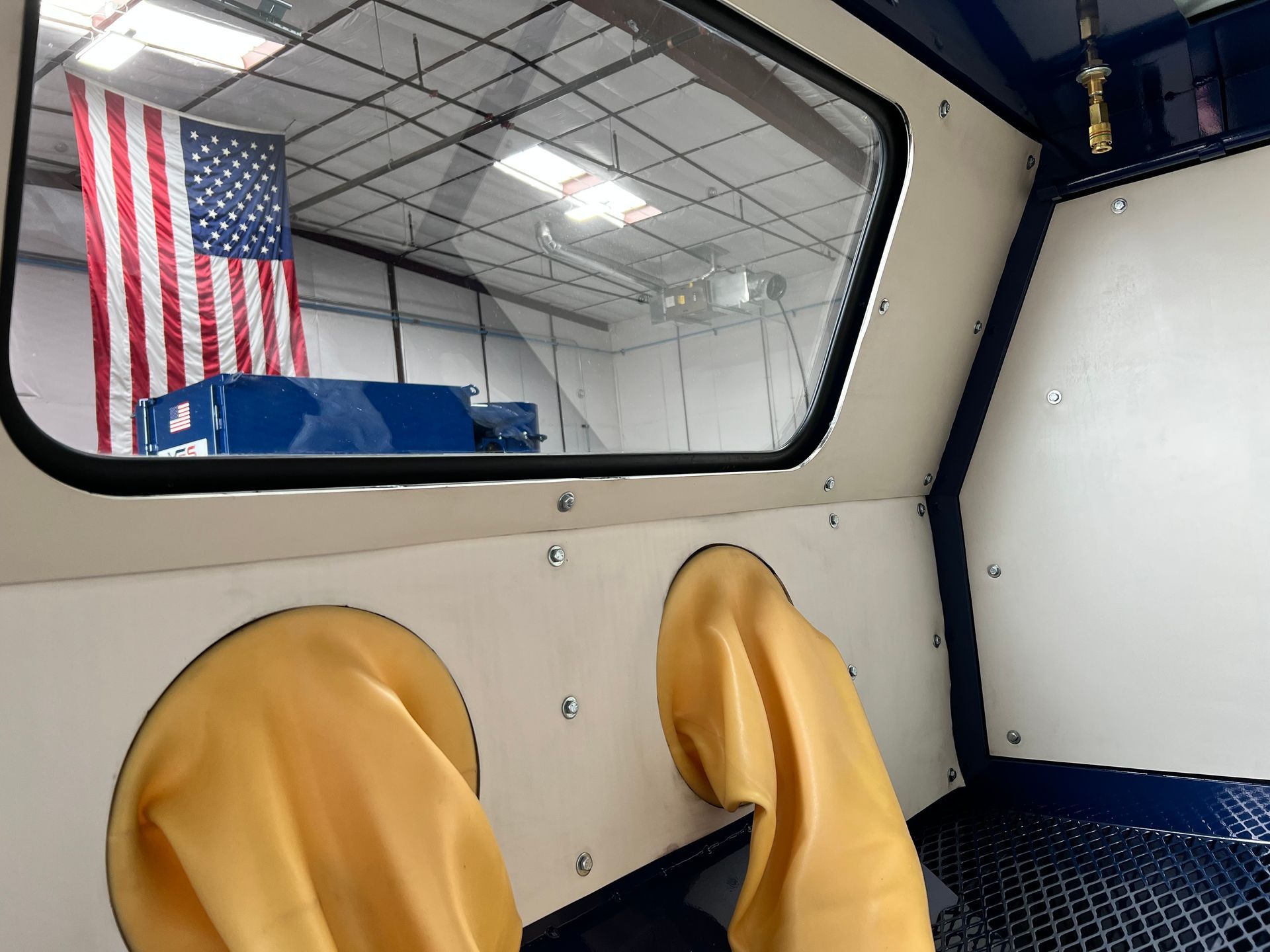 Interior view of a sandblasting booth, with yellow gloves and an American flag visible through the window.