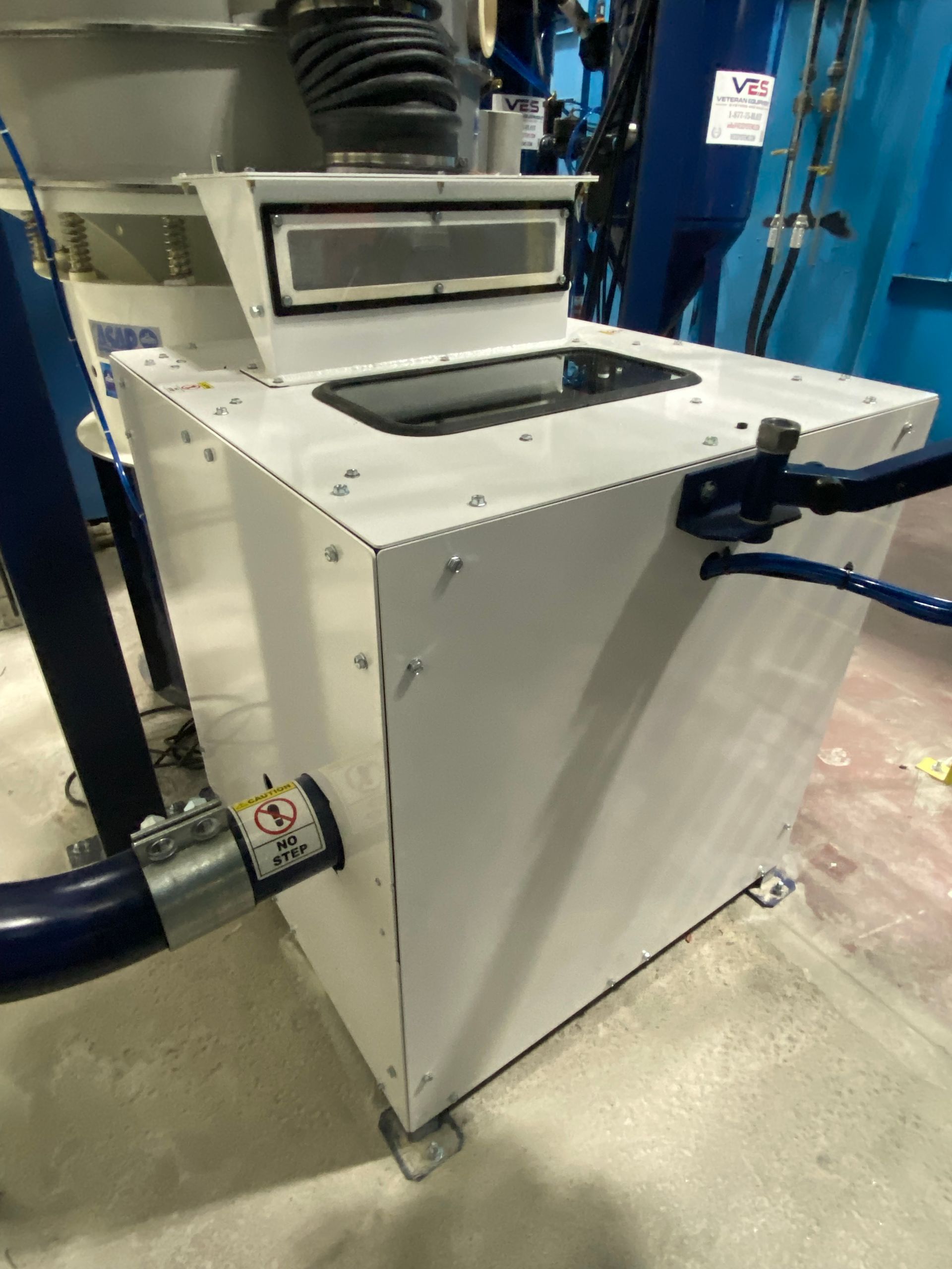 White industrial machine with attached blue hose and handle, in a factory setting.