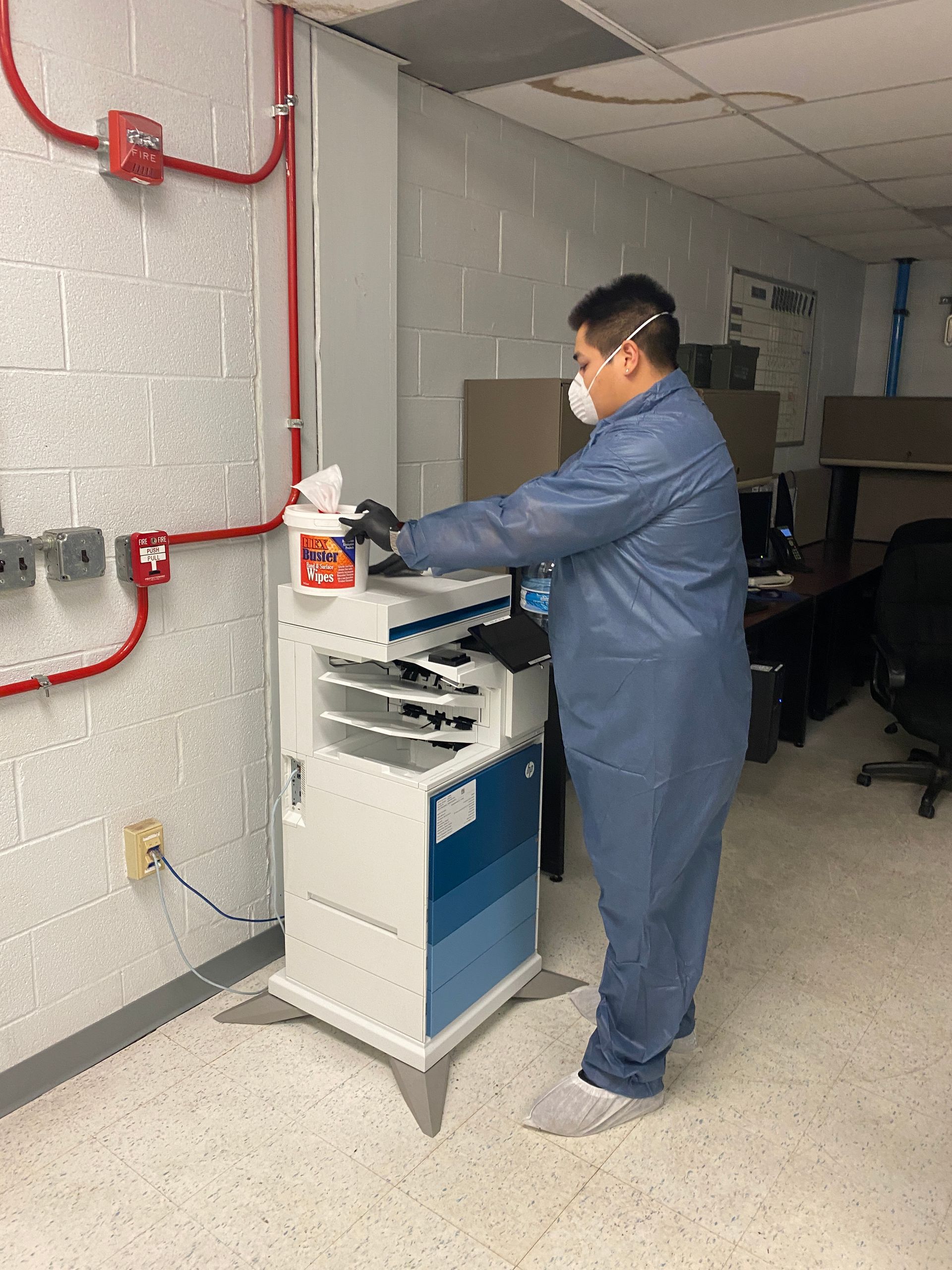 Man in protective gear operates a machine in a lab. He wears a mask, gloves, and a blue jumpsuit.