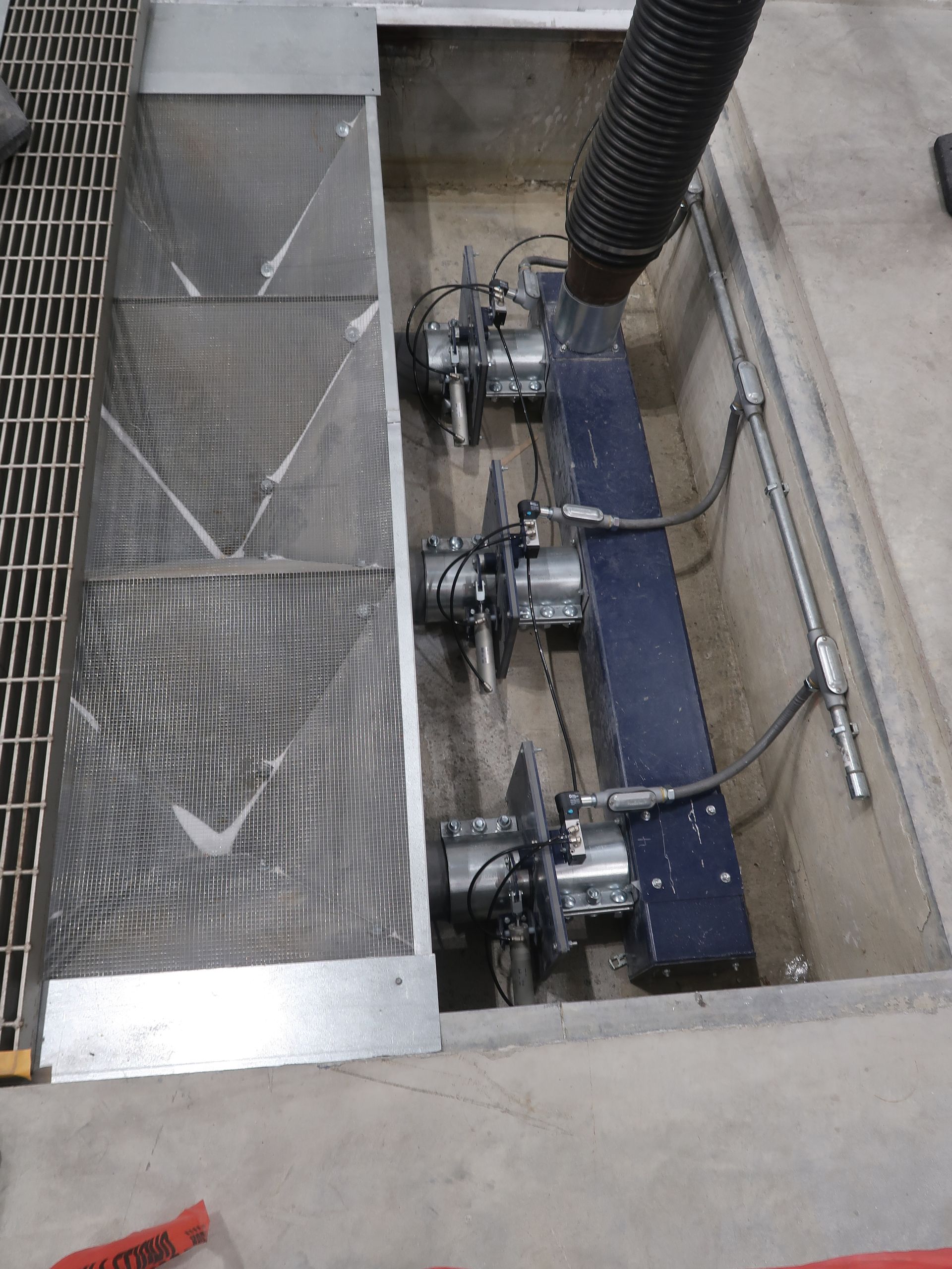 Industrial equipment inside a concrete pit. Metal grates cover part of the opening. Dark blue machinery and piping visible.