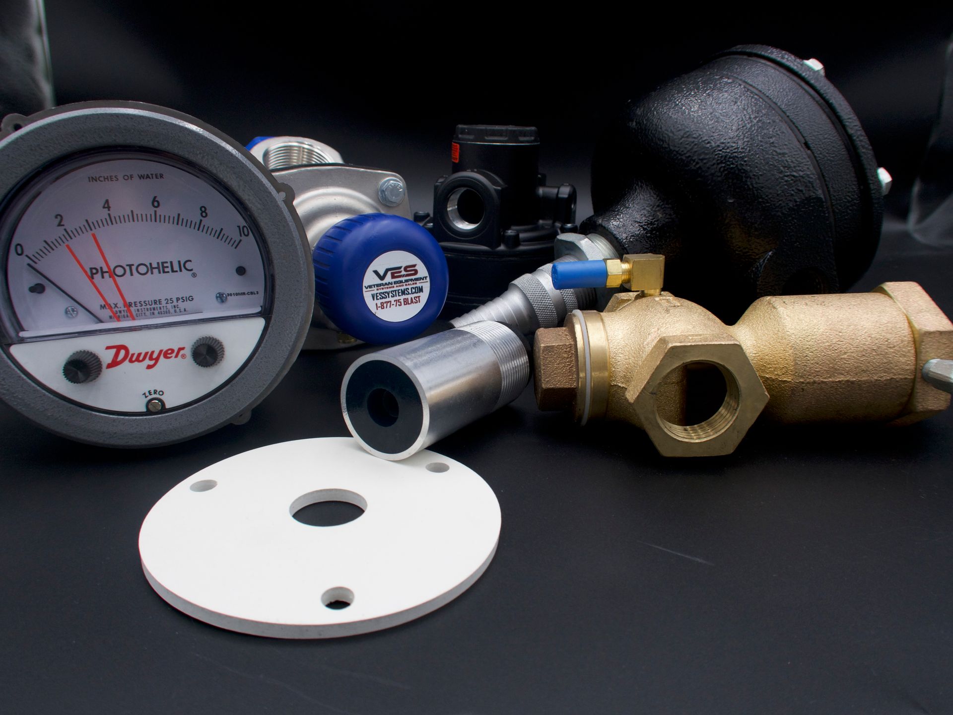 Assorted industrial gauges and valves on a black surface, including a pressure gauge and a brass ball valve.