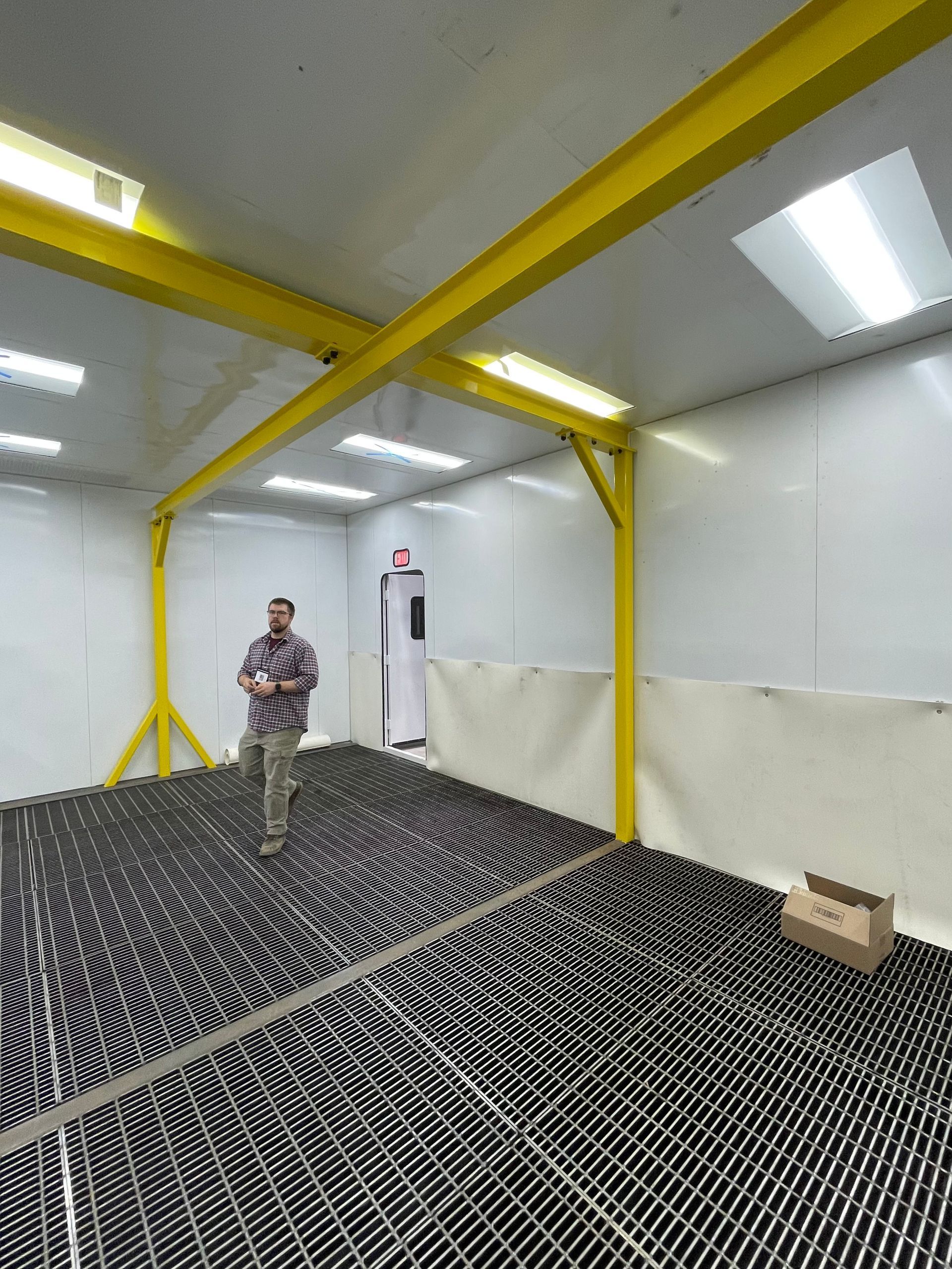 Man standing in a white-walled room with a black grid floor, yellow beams, and a small box.