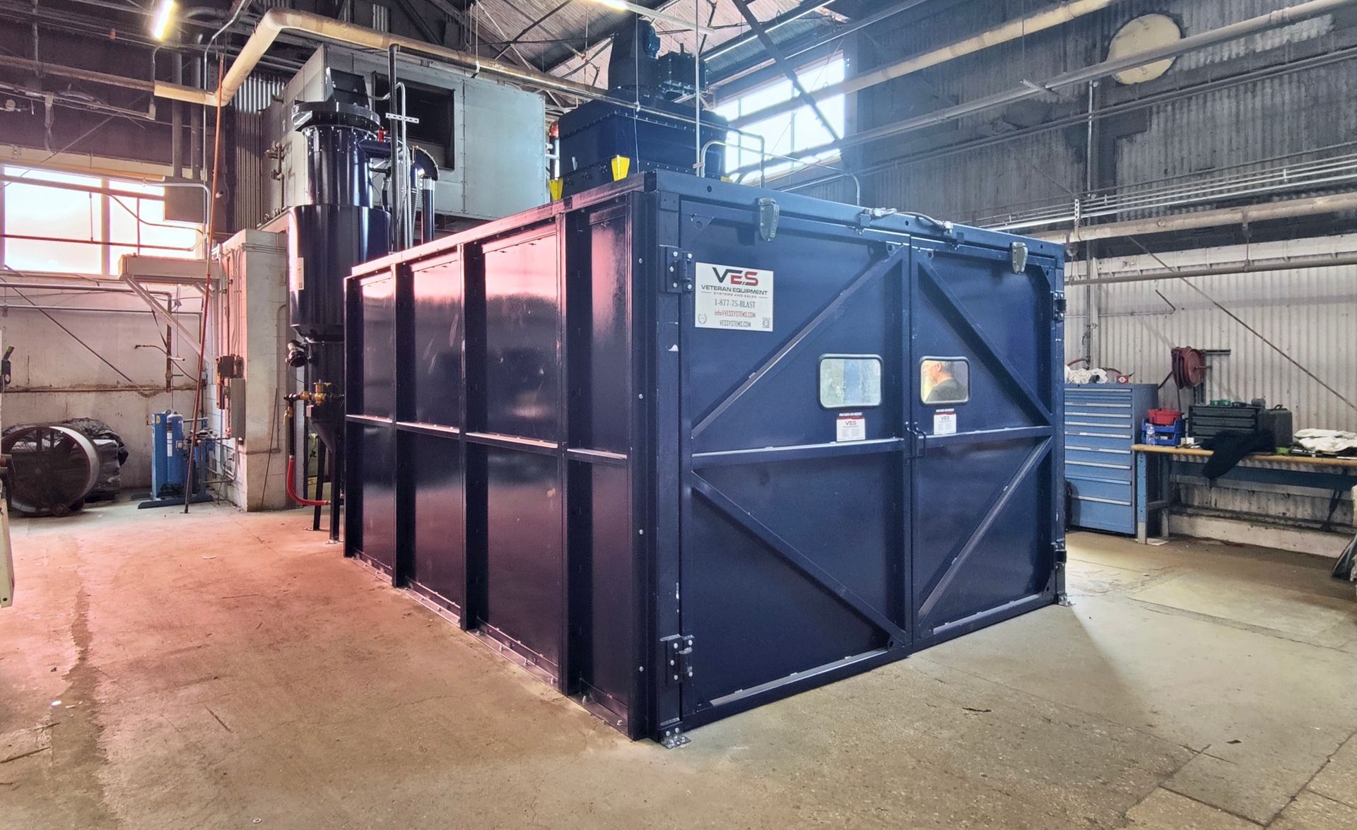 A large, dark-colored industrial container in a factory setting with machinery and pipes.