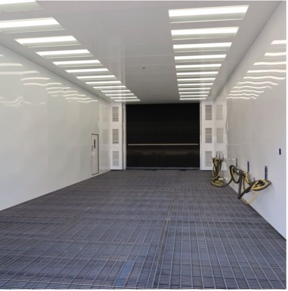 Inside of a white paint booth with a metal grate floor, large black door, and overhead lights.