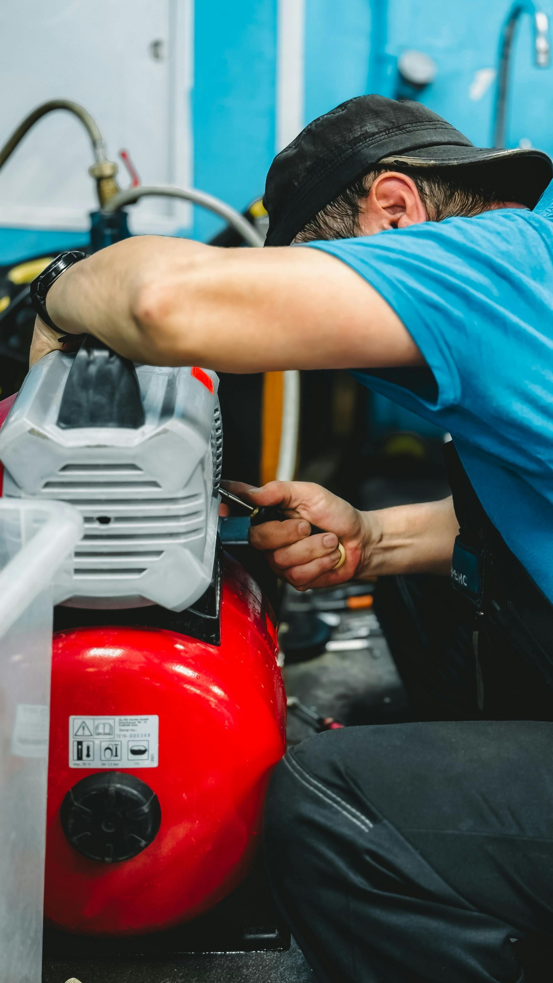Man in blue shirt and black hat fixing red and silver air compressor.