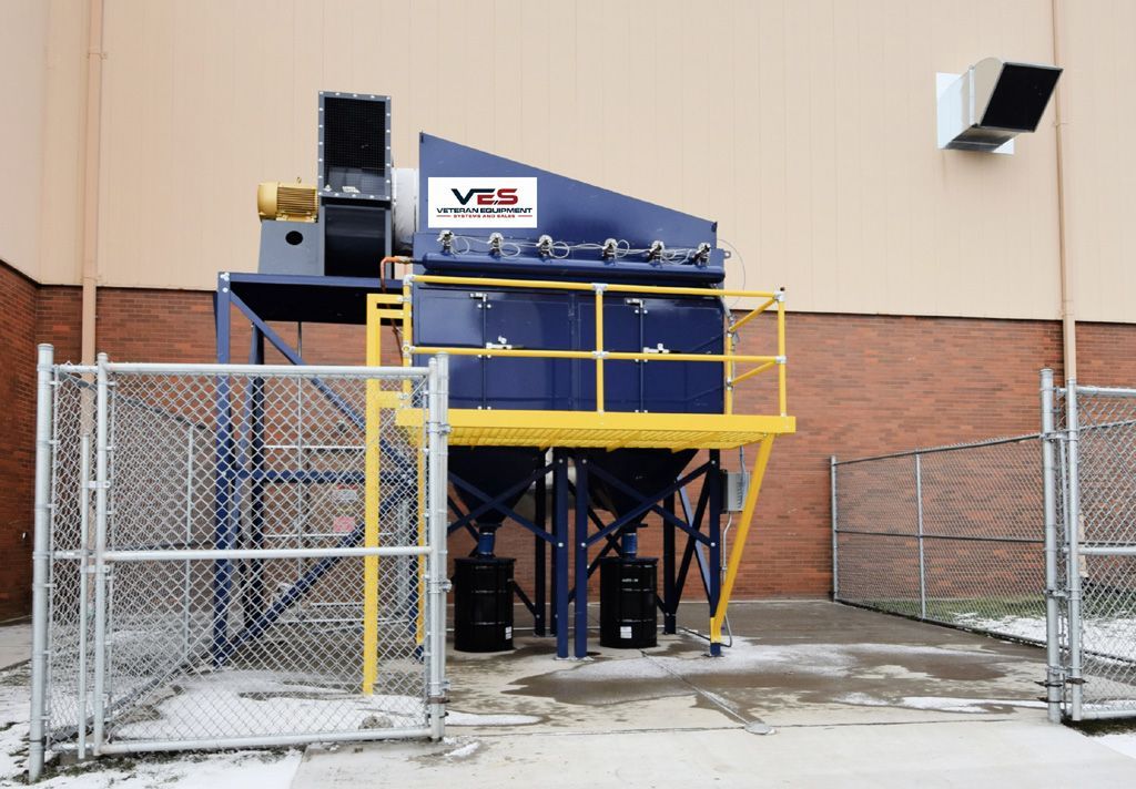 Blue industrial shredder outside a brick building, with a yellow platform and black barrels.