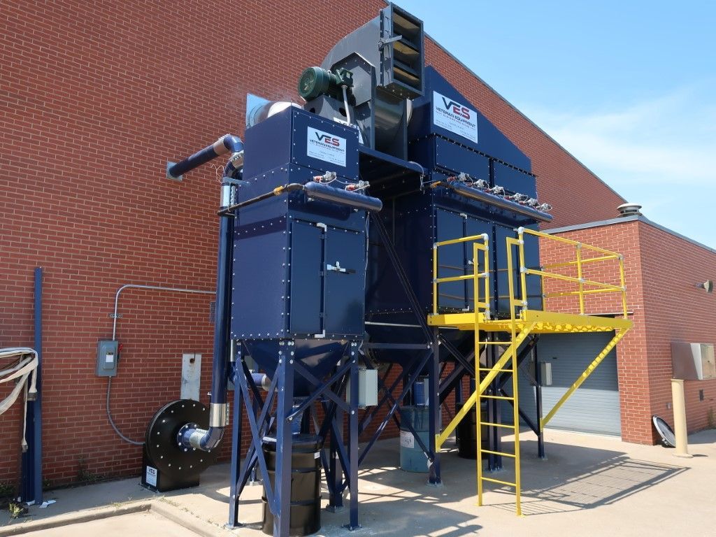 Blue industrial dust collection system outside a brick building with yellow platform and stairs.