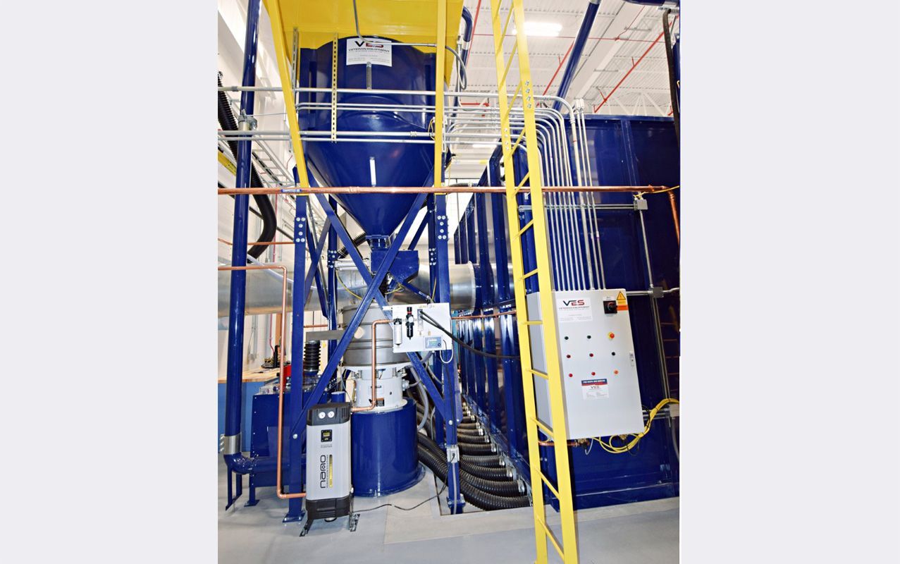 Blue industrial machine with a tall hopper and connected pipes, set in a factory.