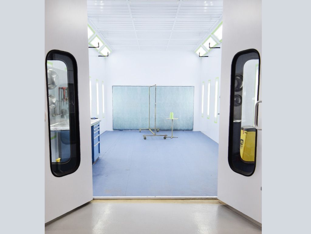 White auto paint booth with open doors, light blue floor, and a folding metal barrier.