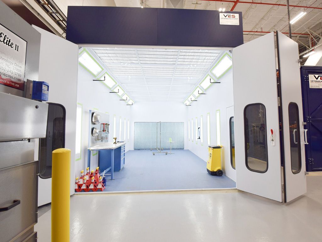 Paint booth interior: white walls, open doors, overhead lights, yellow safety pole, blue table.