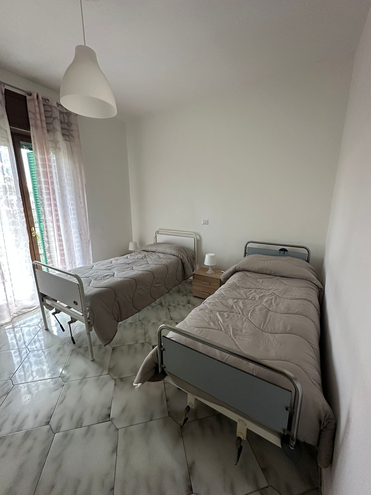 Due letti d'ospedale in una stanza luminosa con pareti bianche, pavimento a motivi e tende trasparenti.