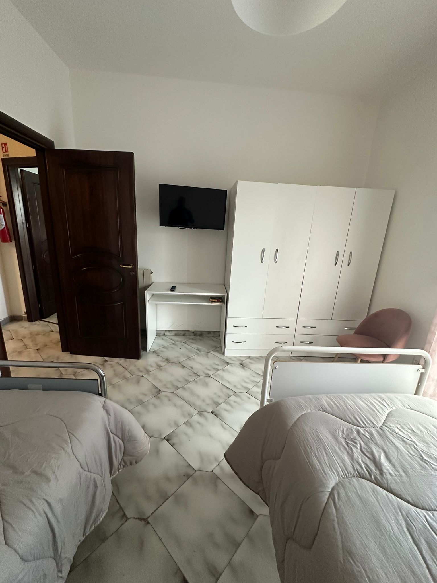 Camera da letto con due letti, armadio, scrivania e TV; biancheria da letto grigio chiaro, pavimento effetto marmo.