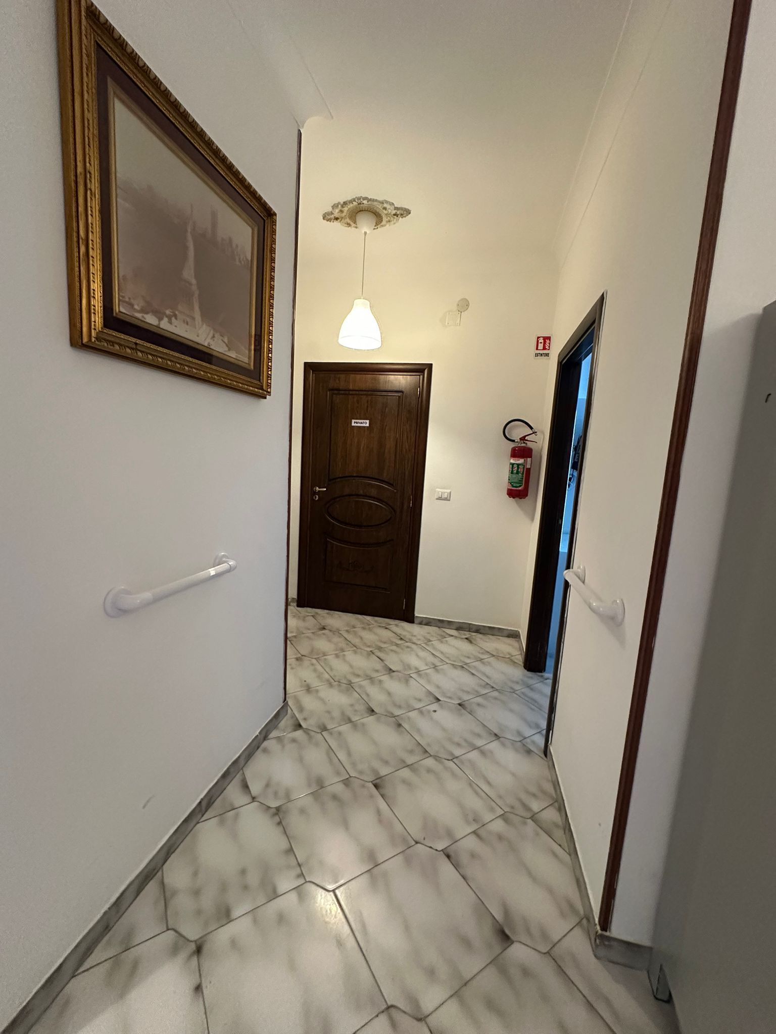 Corridoio con pavimento in piastrelle di marmo, quadri incorniciati, porta marrone e porta aperta.