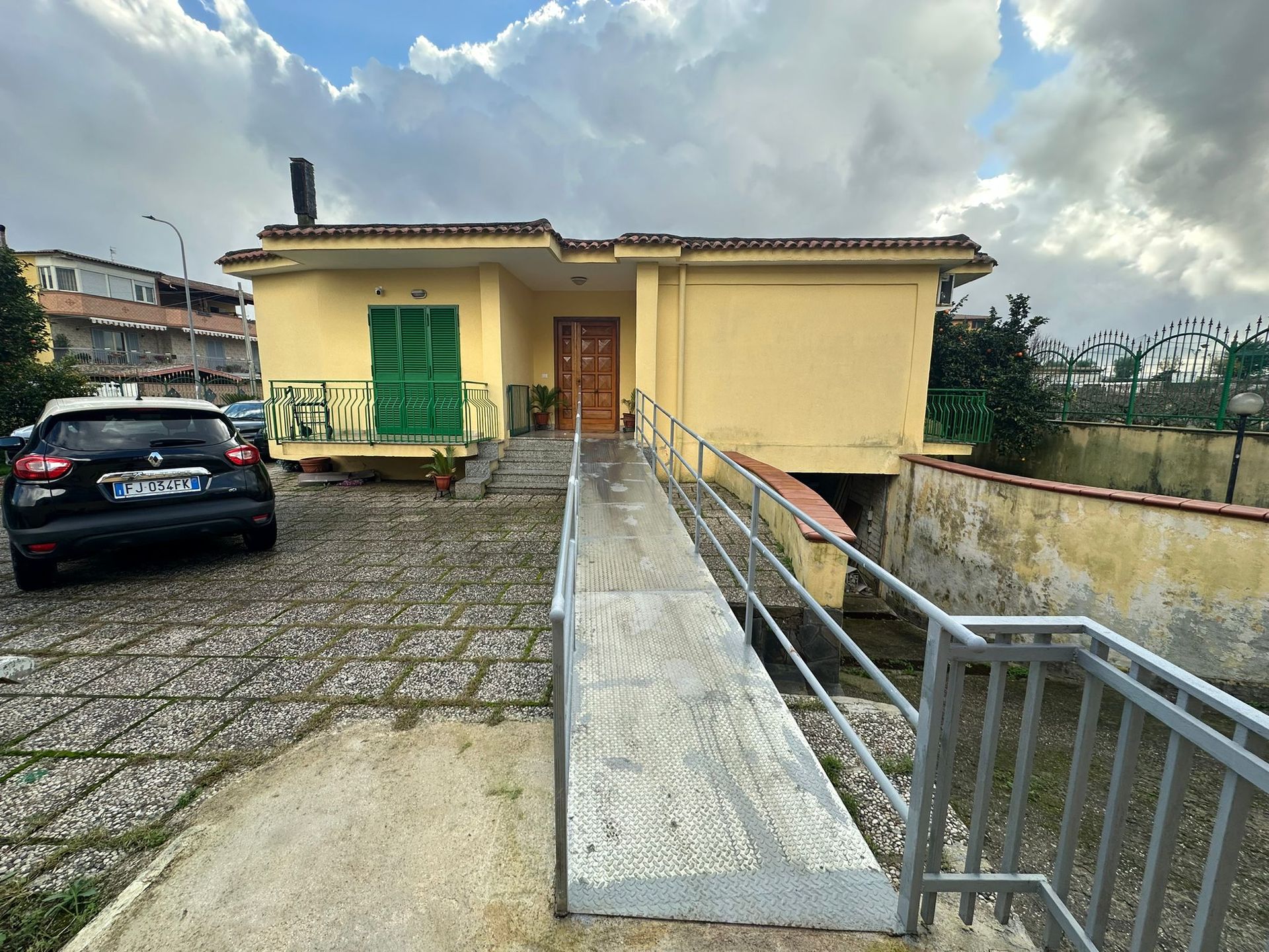 Edificio giallo con rampa, auto parcheggiata davanti. Persiane verdi, porta marrone. Cielo nuvoloso.