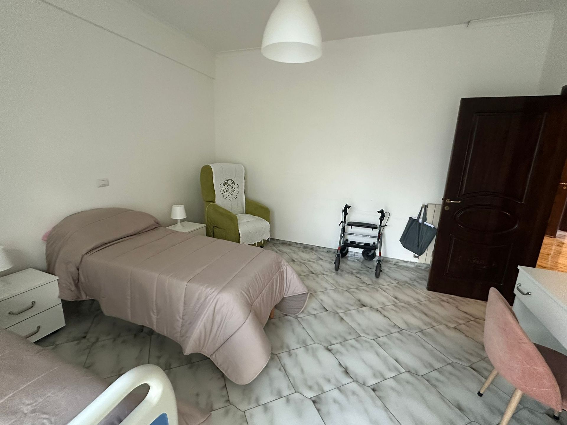 Camera da letto con due letti, poltrona verde, deambulatore e pavimento in marmo bianco.