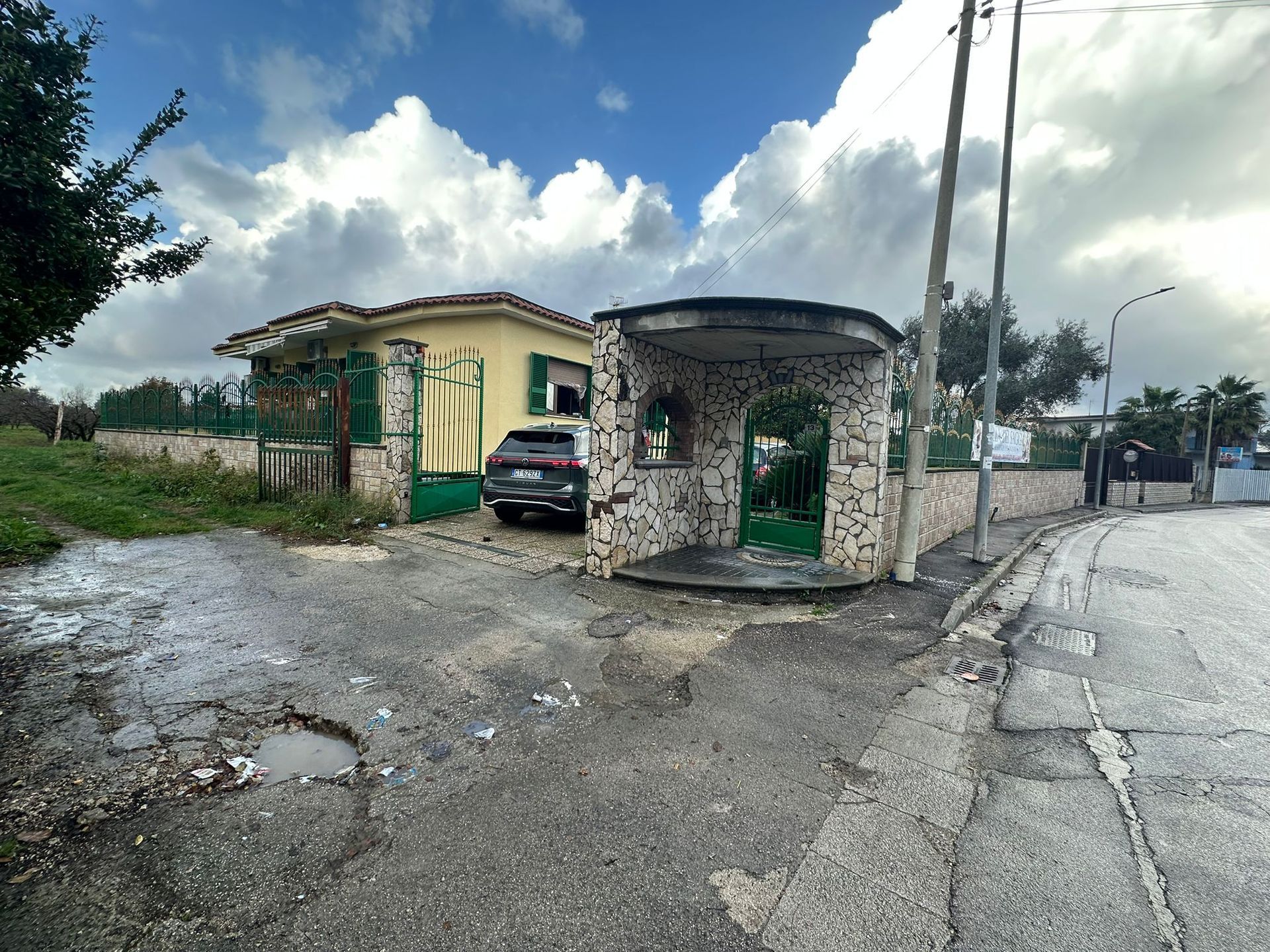 Un'auto parcheggiata fuori da una piccola casa color sabbia con un cancello verde e una strada asfaltata irregolare. Cielo nuvoloso.