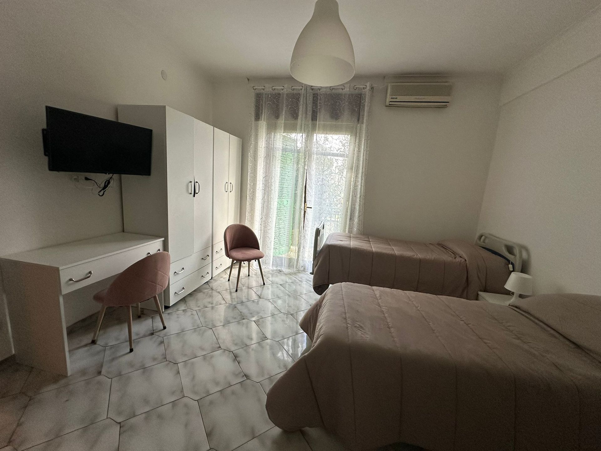 Camera da letto con due letti singoli, scrivania con sedia, armadio e finestra con tende trasparenti.