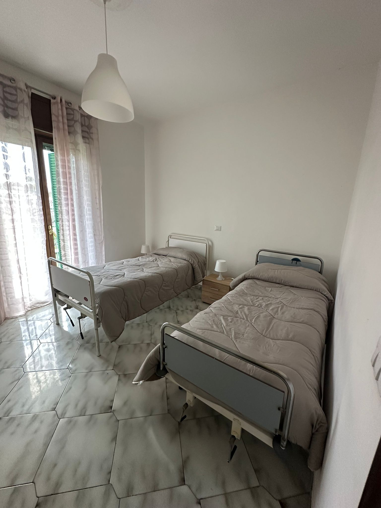 Due letti singoli in una stanza con una finestra, pareti bianche e una lampada sospesa.