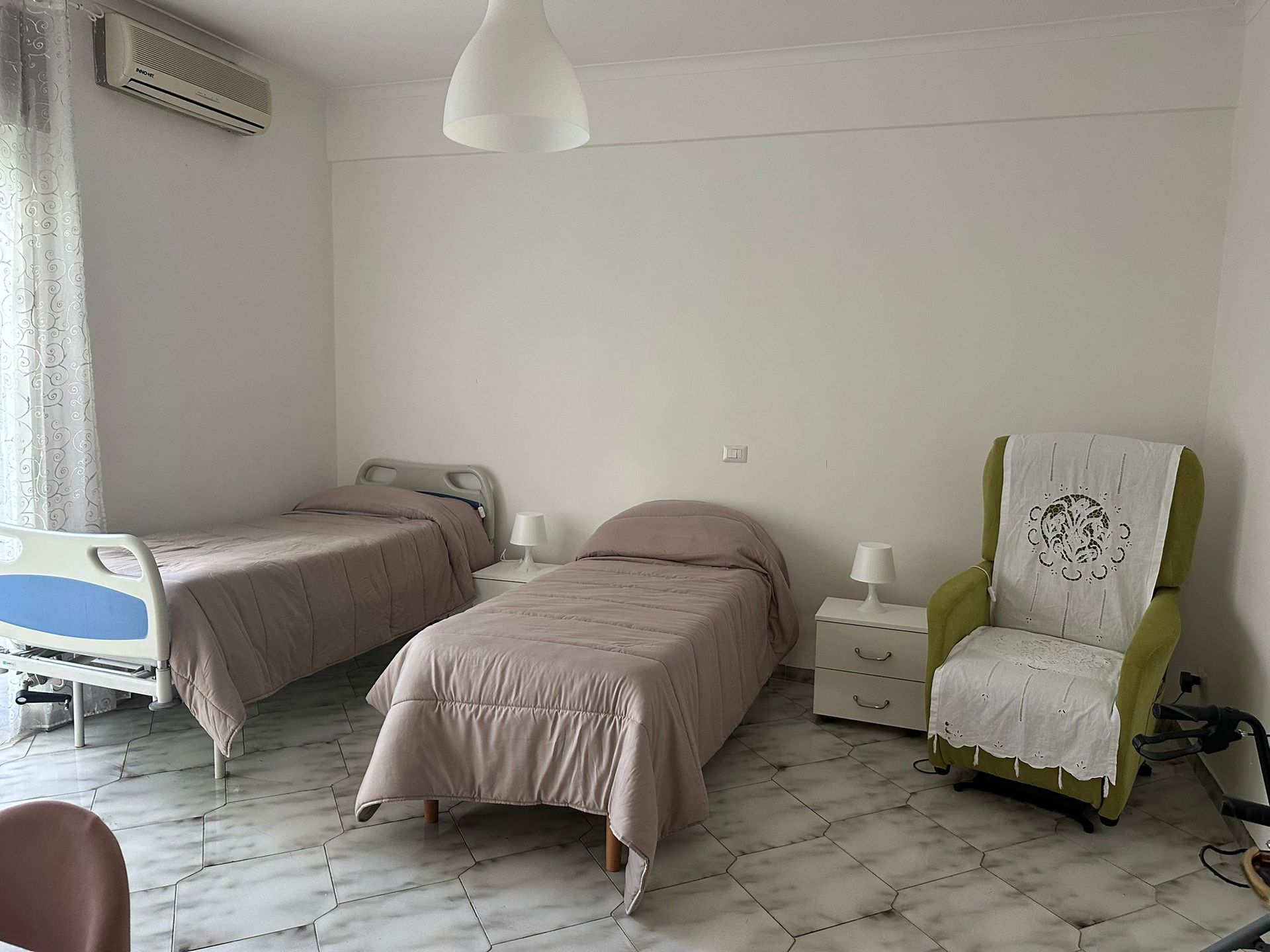 Camera da letto con due letti, una sedia e un comodino. Pareti e pavimento chiari, luce naturale.