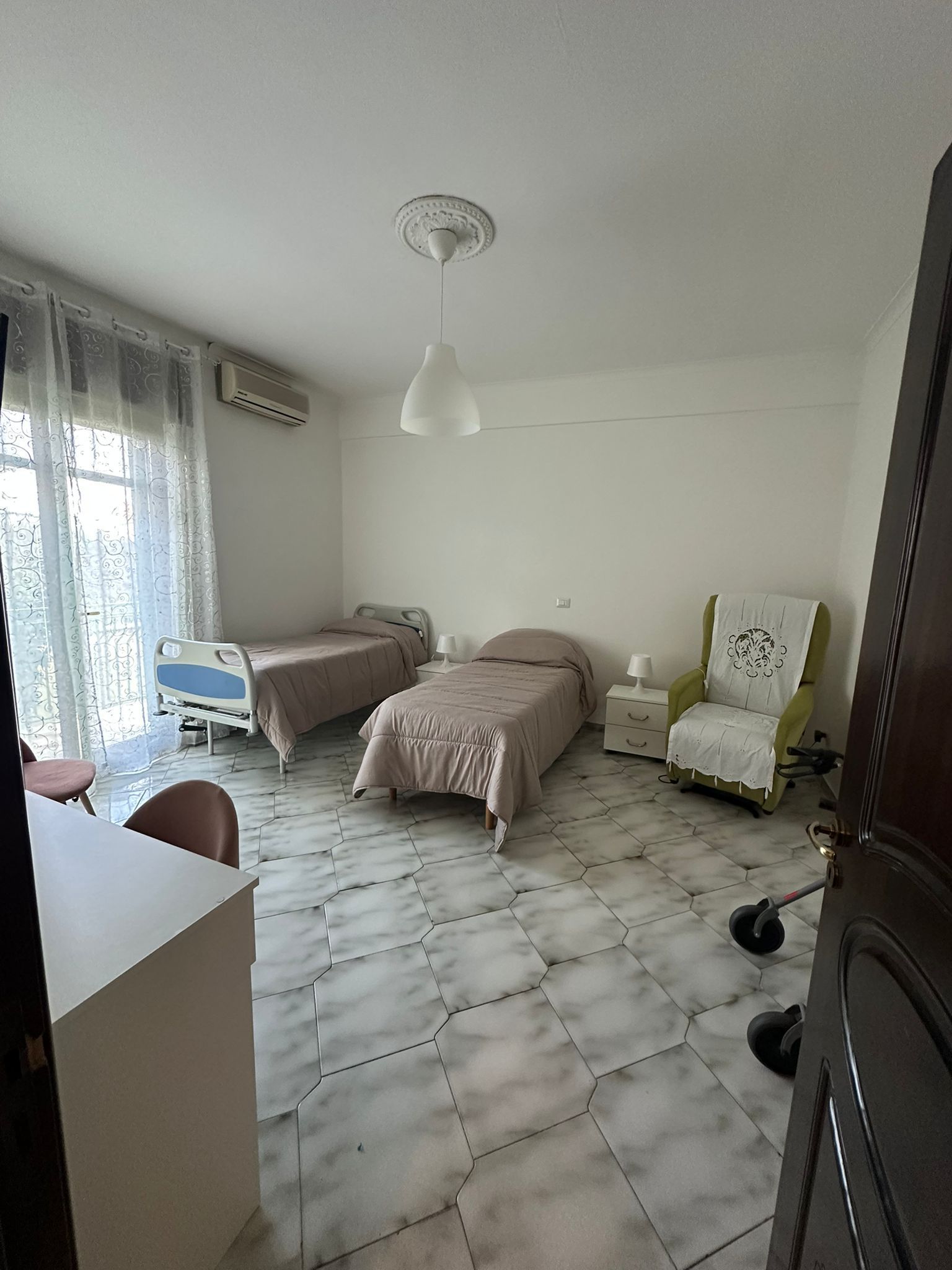 Due letti, una poltrona e un armadio in una stanza con pavimento e pareti chiari; in primo piano c'è una porta.