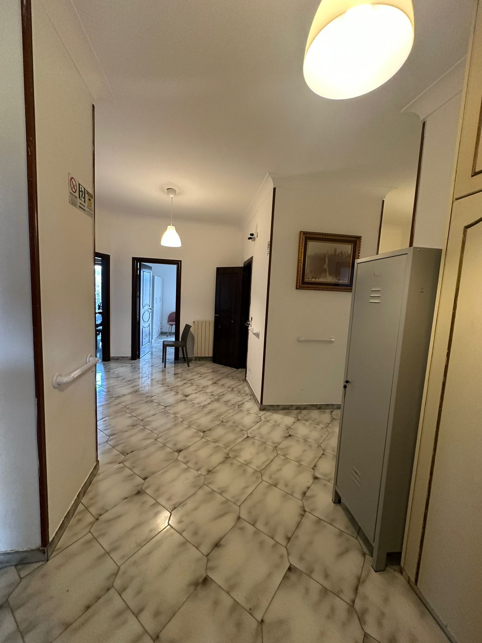 Corridoio con pavimento piastrellato, più porte, una lampada e un mobile grigio.