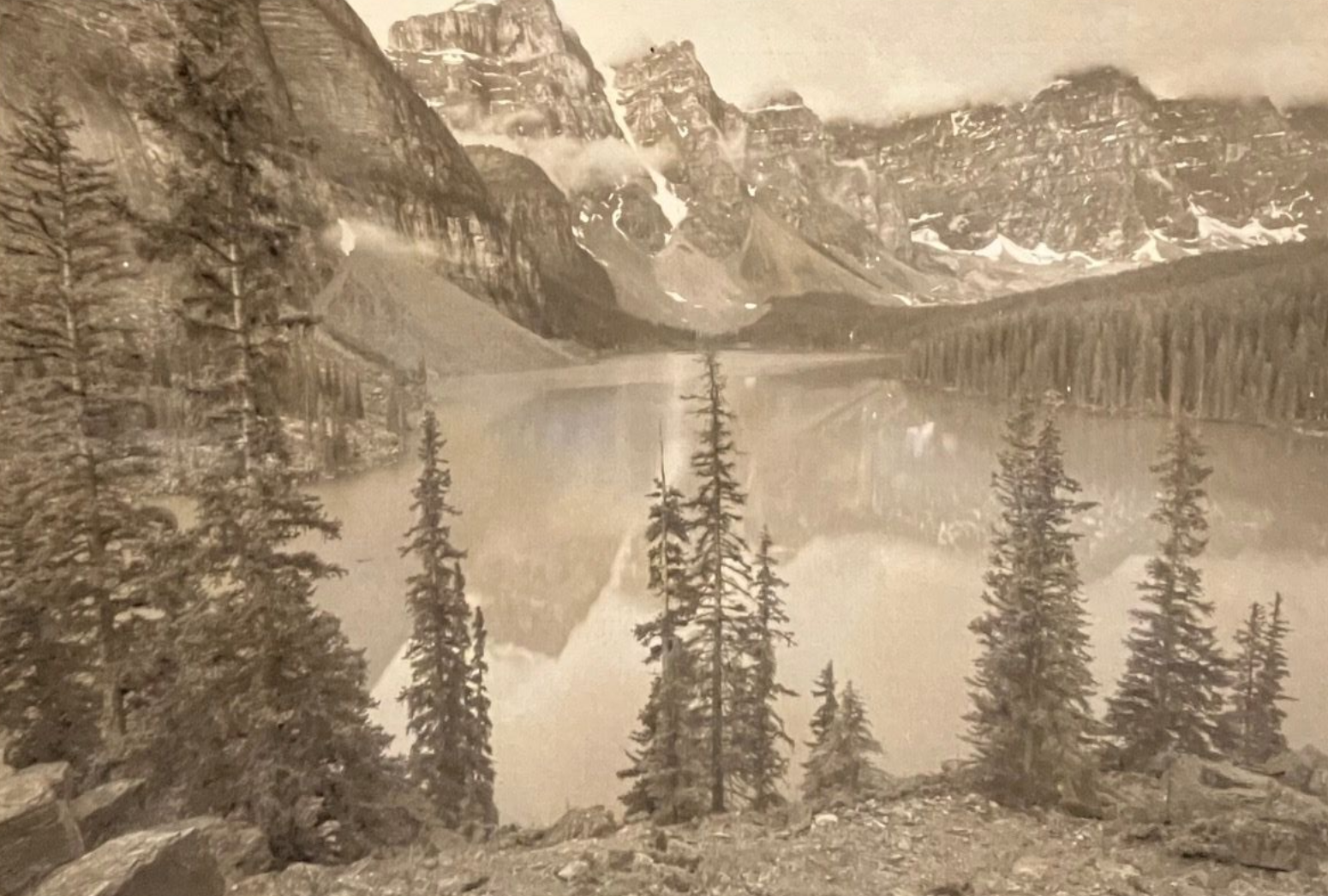 Mountains reflected in a calm lake, framed by pine trees, sepia-toned.