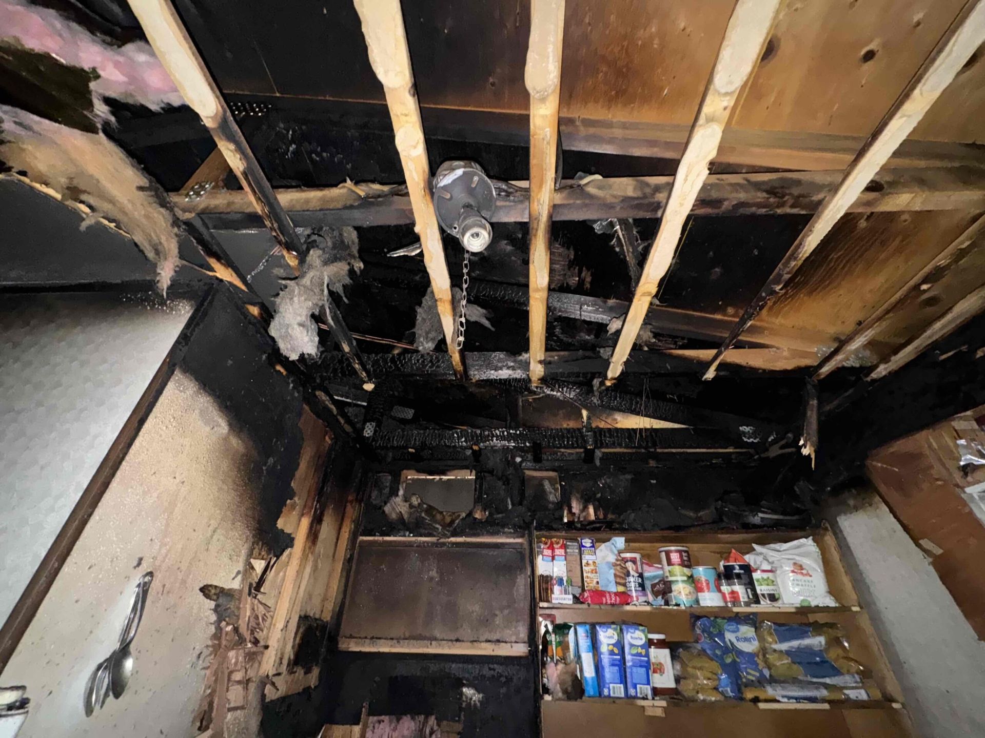 Interior view of a fire-damaged kitchen; charred ceiling, cabinets, and contents visible.
