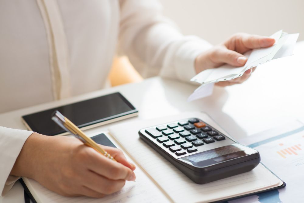 Person calculating expenses, holding receipts, with a calculator, notepad, and smartphone on a desk.