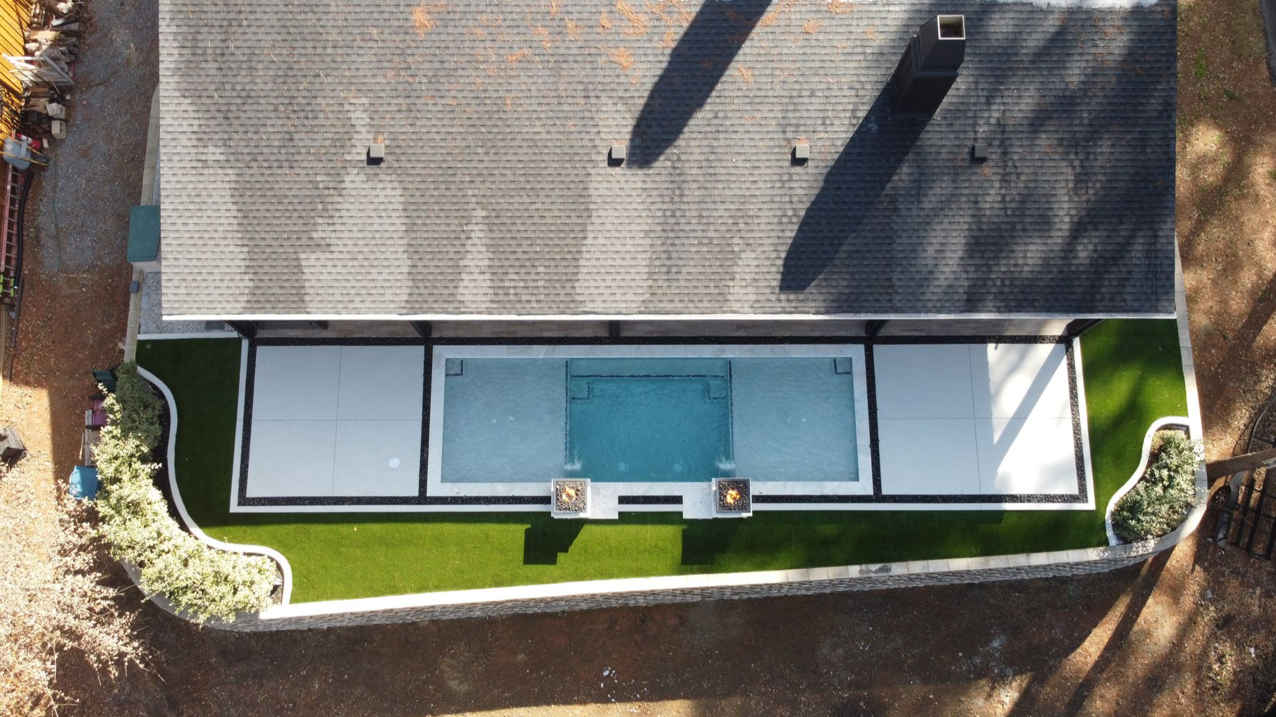 Overhead view of a house with a rectangular pool and artificial turf lawn. Grey roof, white and black patio.