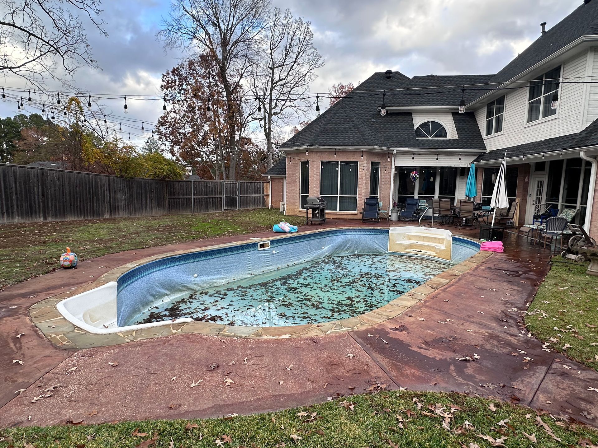 Backyard with a pool covered in debris, next to a brick house with a dark roof on an overcast day.