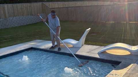 A person using a long-handled pool skimmer to clean a swimming pool in a backyard at sunset.