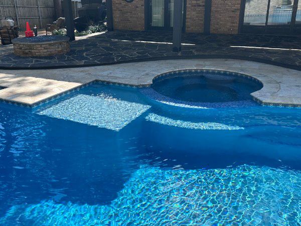 A bright blue backyard swimming pool featuring a built-in circular spa and a shallow sun shelf with stone decking.