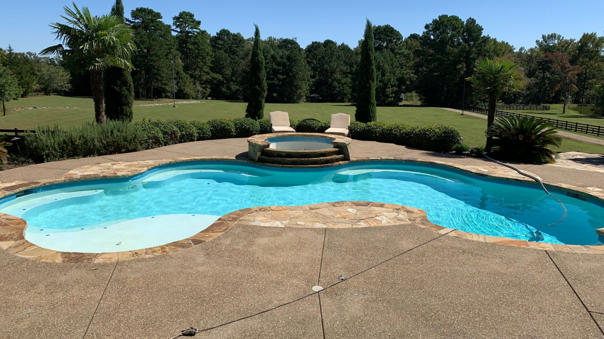 Swimming pool and hot tub surrounded by stone patio with backyard view of trees.