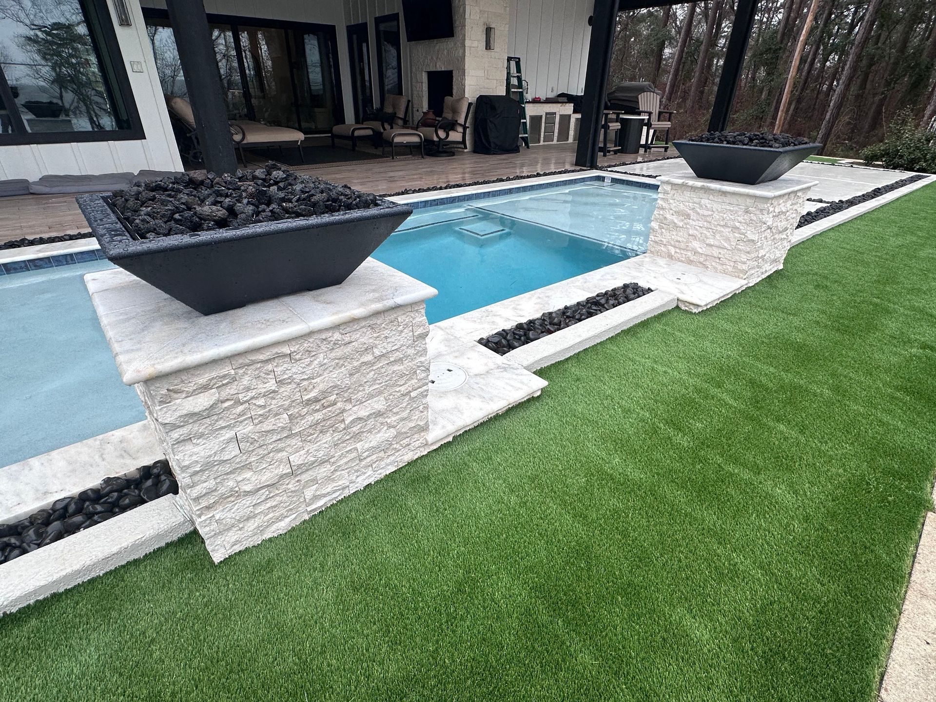 Outdoor pool with fire pit, stone pillars, black rock accents, and green turf.