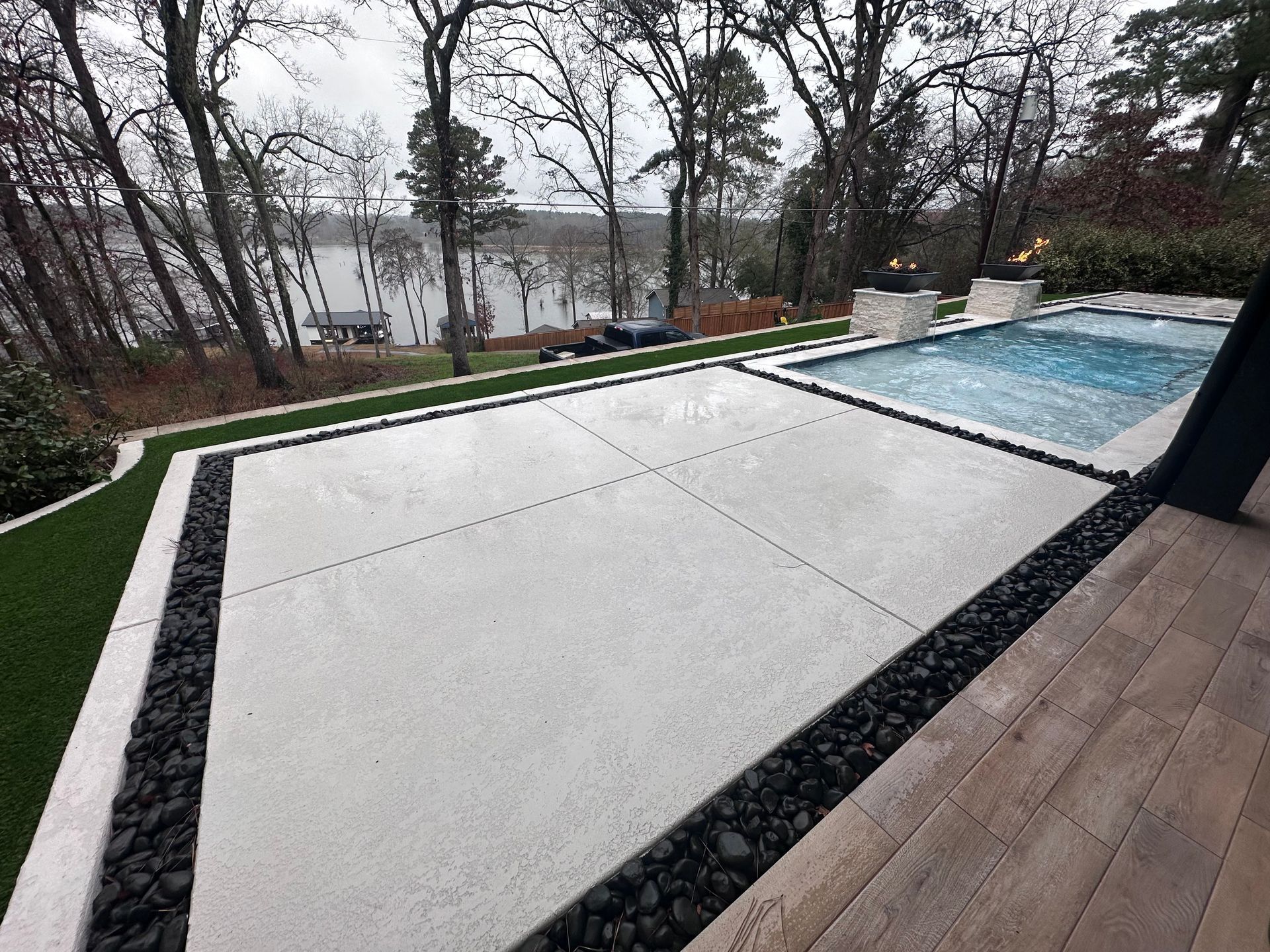 Concrete patio with pool, black river rock border, artificial turf, and a lake view.