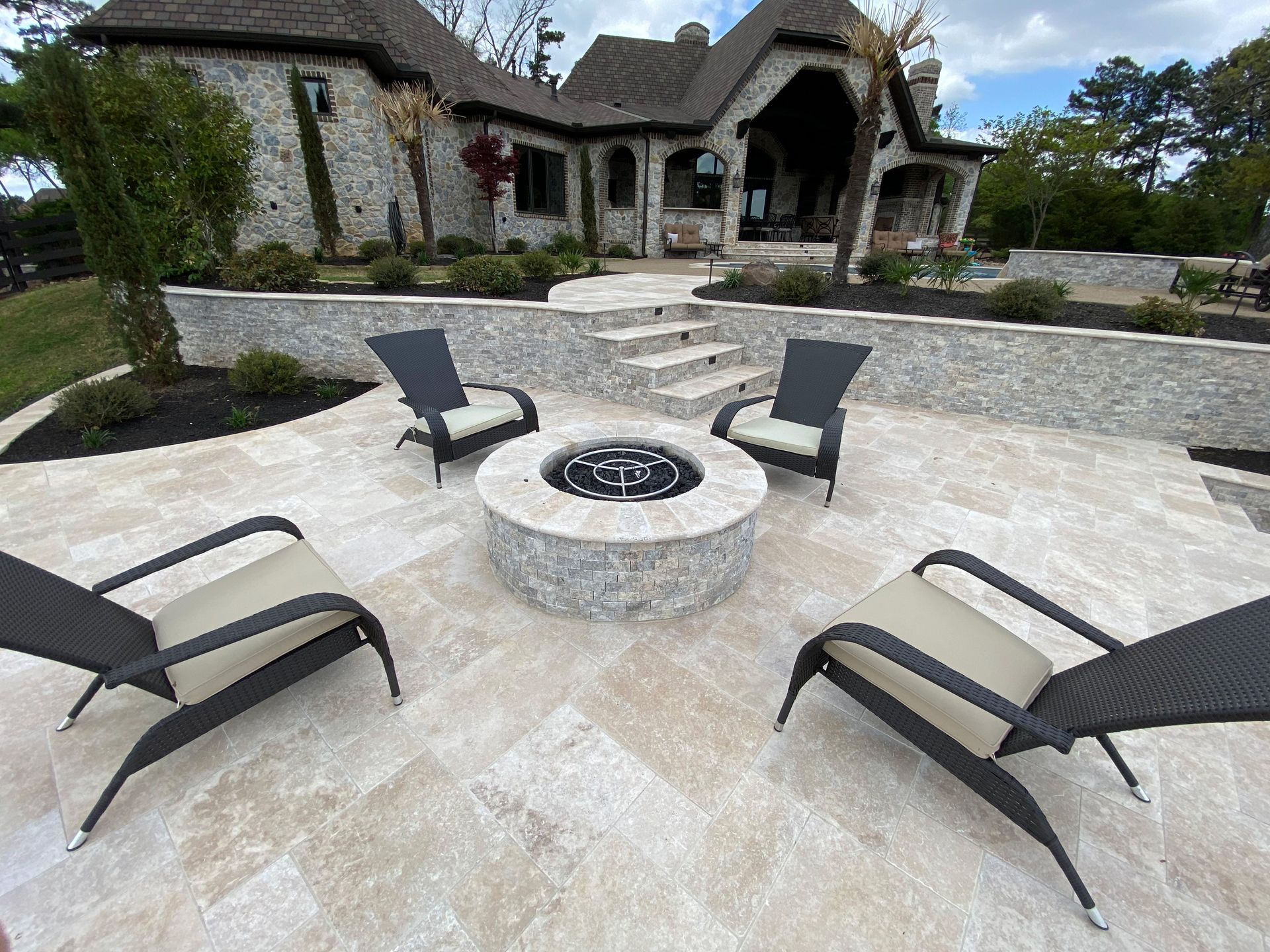 Outdoor patio with stone fire pit and four modern chairs, in front of a large stone house.