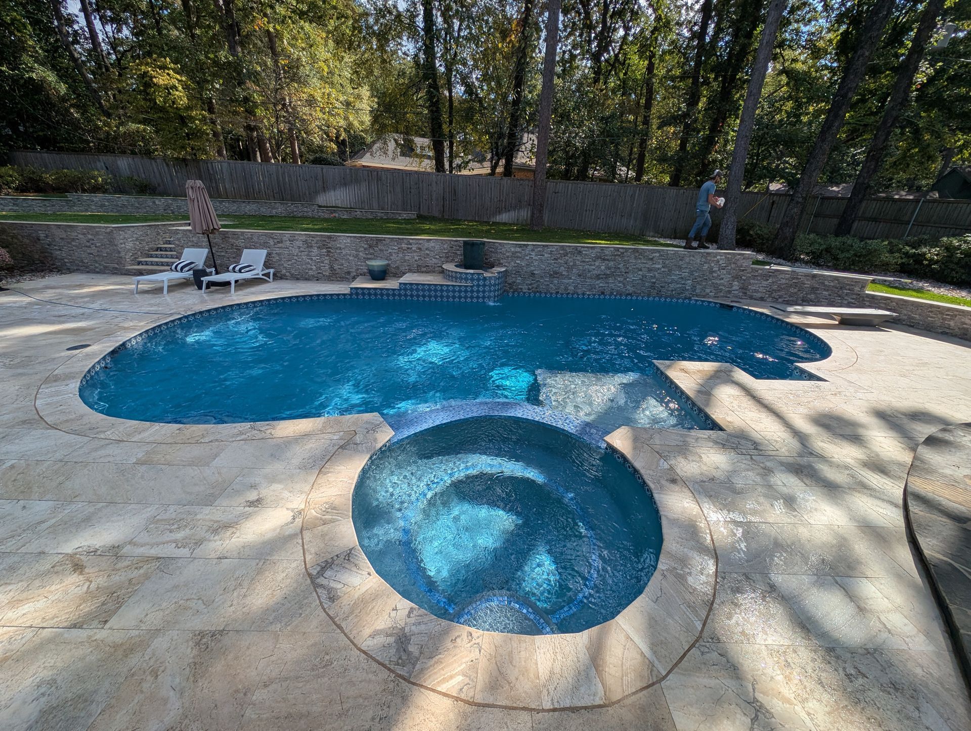 Pool with blue water and adjacent spa in a backyard setting. Trees in the background.