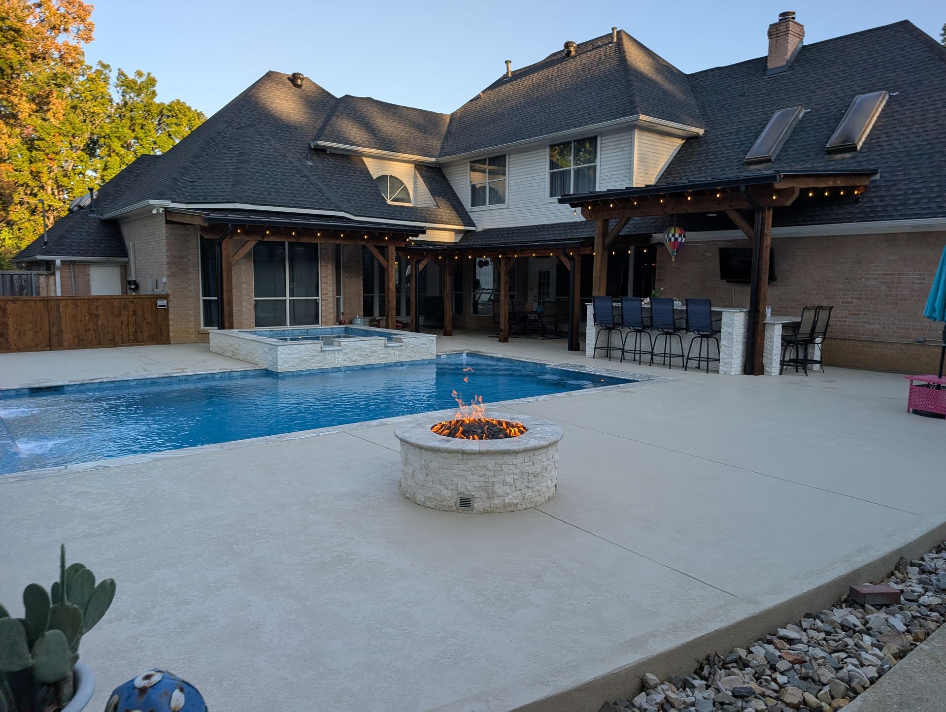Backyard with pool, hot tub, and fire pit. Pergola, bar seating, and a large house in the background.