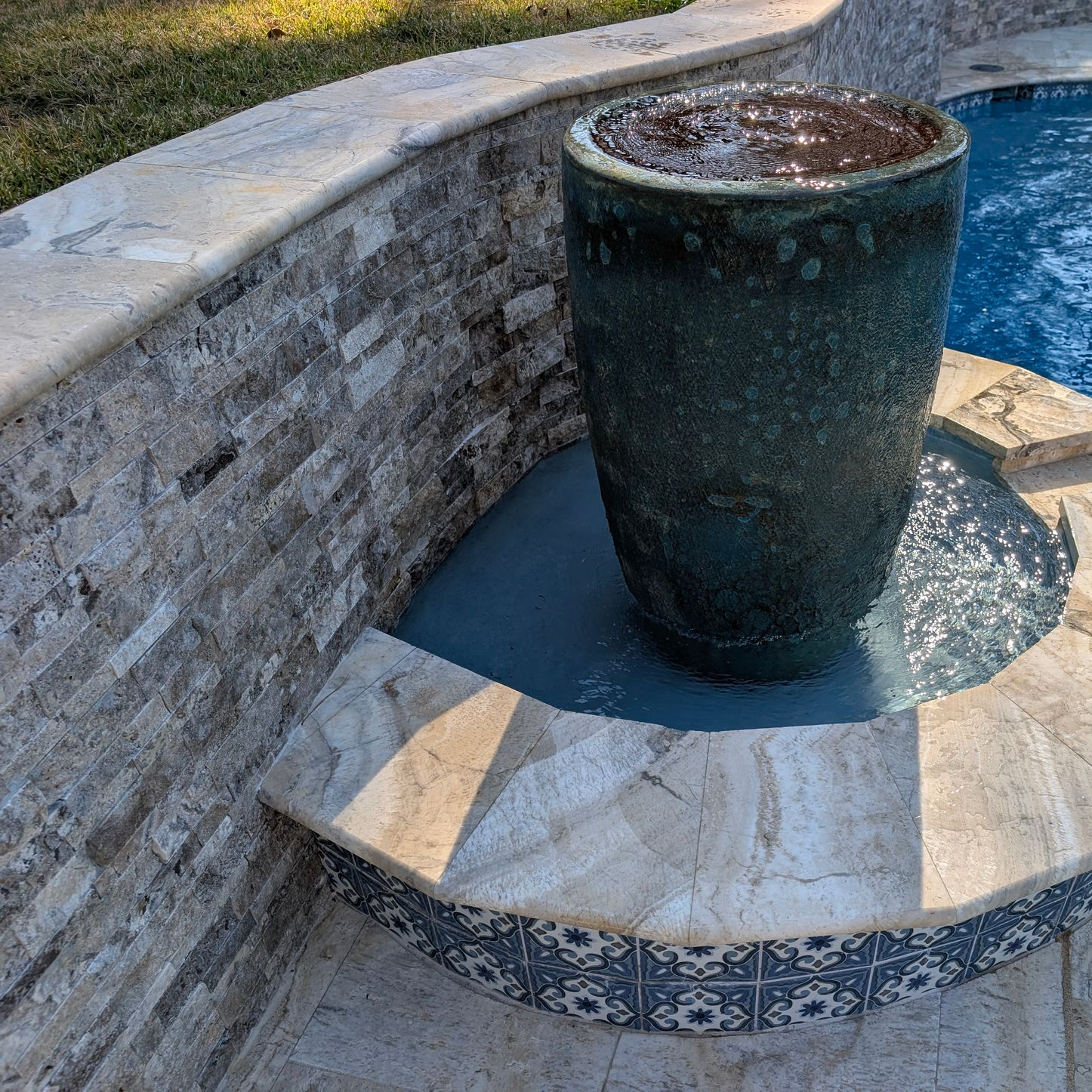 A dark, textured fountain pot sits in a tiled pool feature against a stone retaining wall.