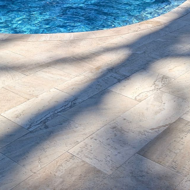 A curved pool edge lined with light-colored, textured stone pavers, partially shaded by palm tree fronds.