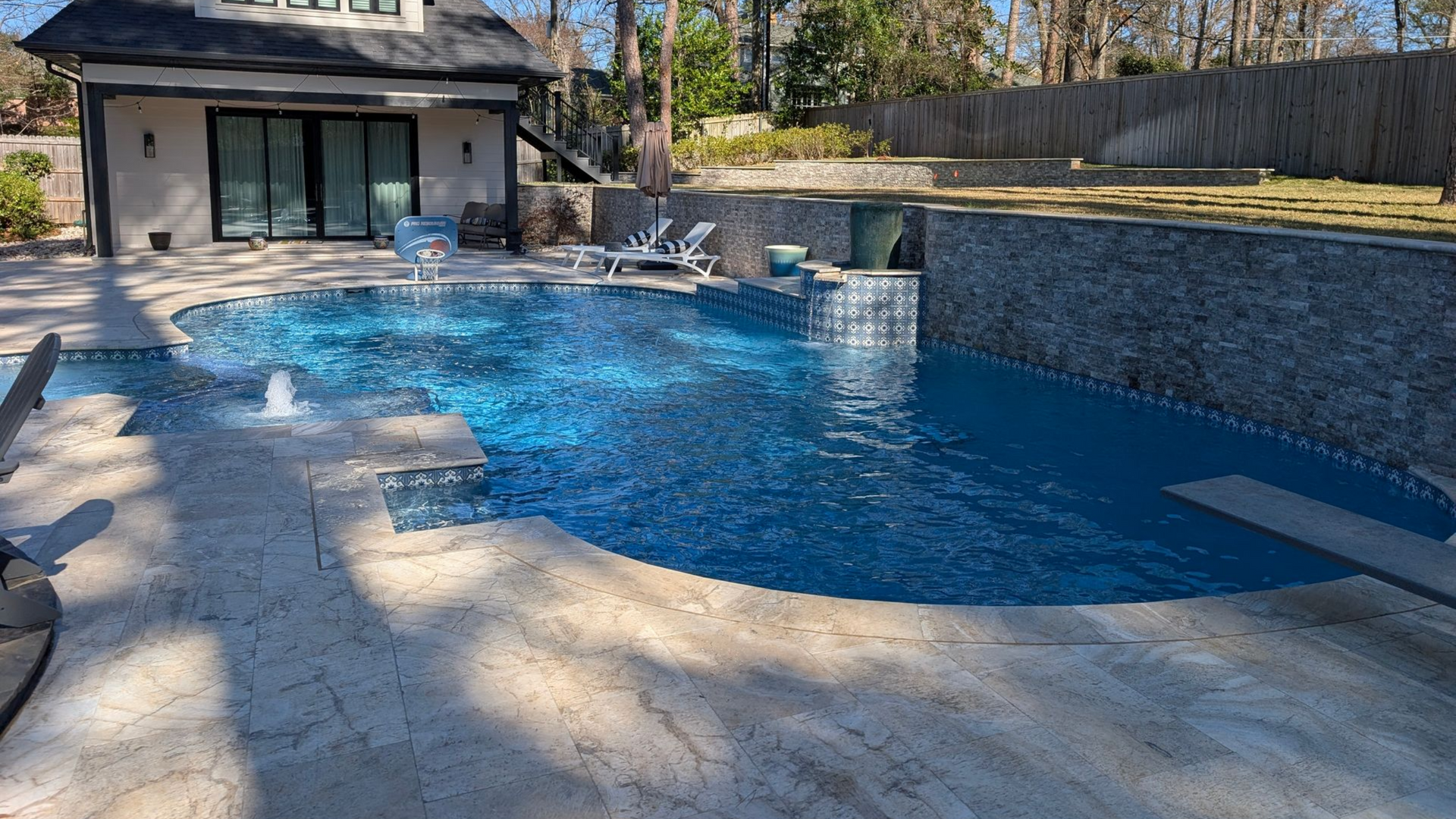 A backyard swimming pool with fountains, a diving board, and a waterfall feature, surrounded by beige stone.