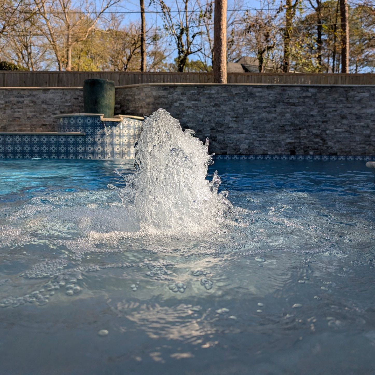 A fountain shoots water into a swimming pool with a stone wall and trees in the background.