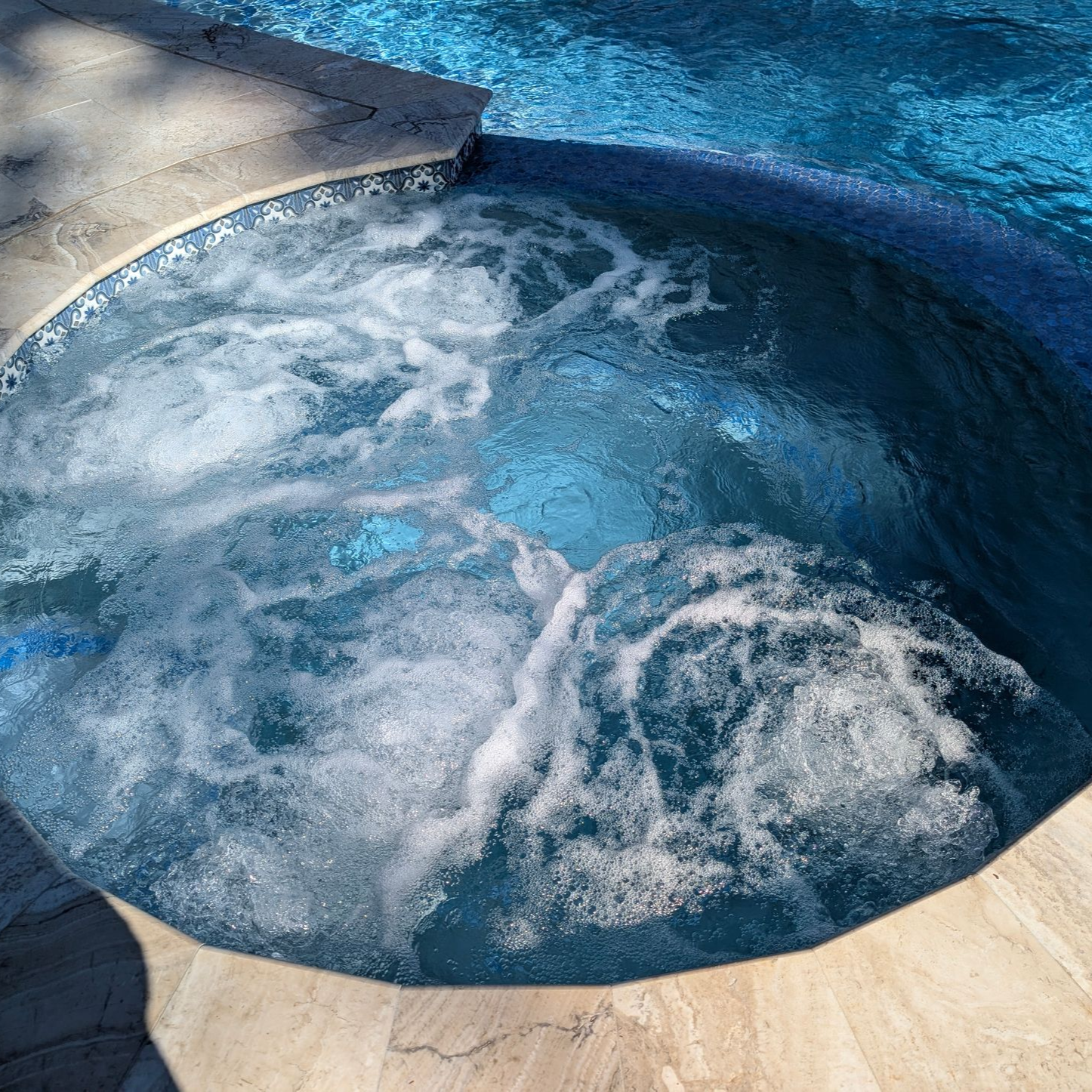 A bubbling circular spa with blue water surrounded by beige stone coping, connected to a larger swimming pool.