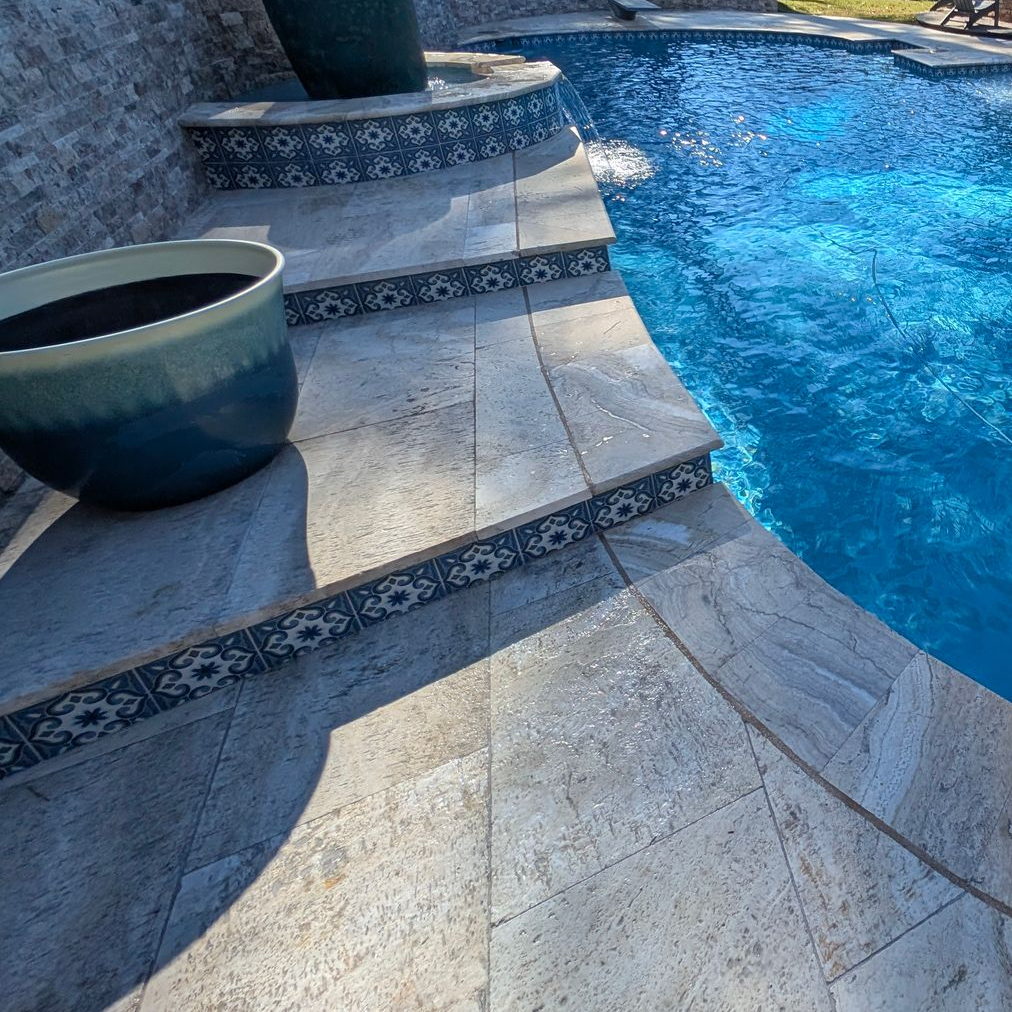 Stone pool deck with decorative tiled step risers leading into a bright blue swimming pool, featuring large teal planters.