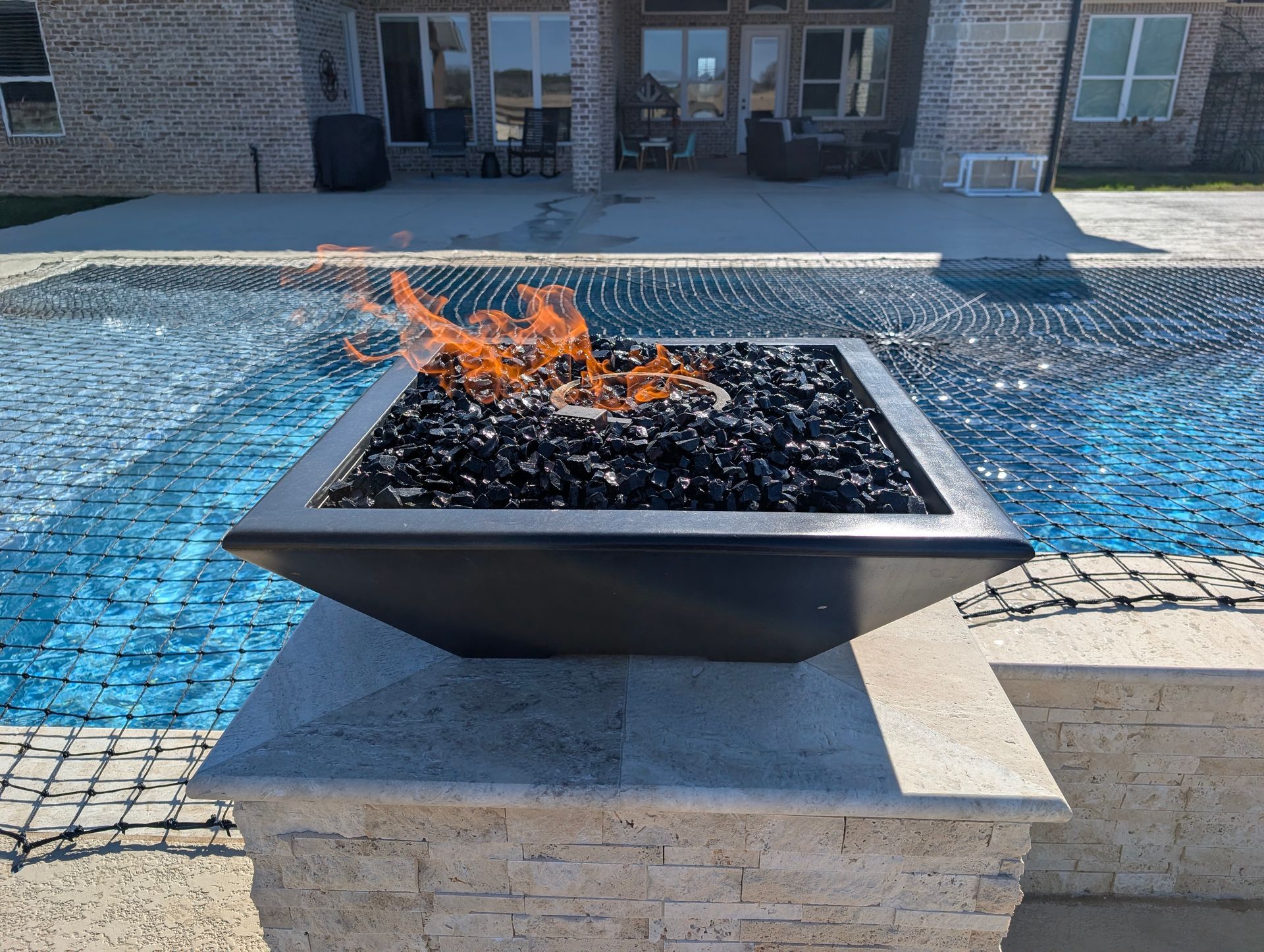 A square fire pit with flames, filled with black glass, sits beside a pool.