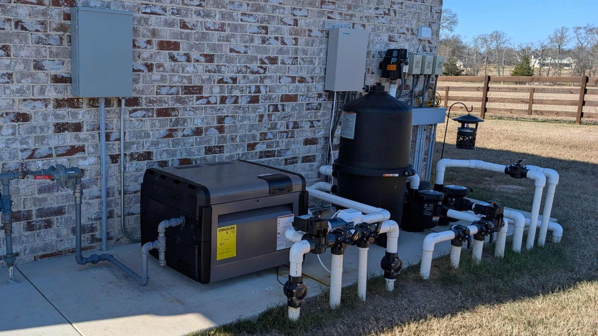 Outdoor pool equipment against a brick wall: heater, filter, pump, control panels, and white PVC pipes.