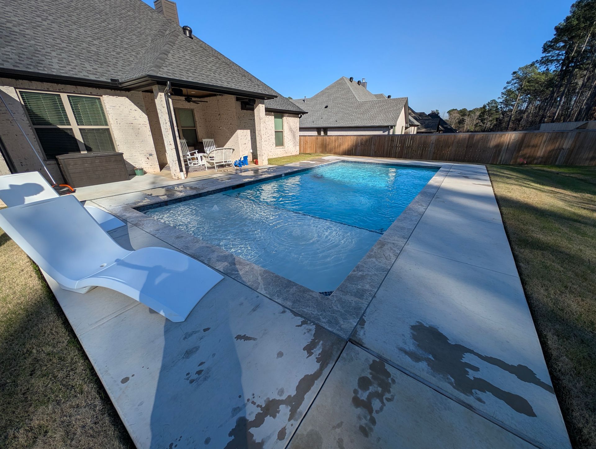 Backyard swimming pool beside a stone patio, with a white lounge chair and house in sunlight