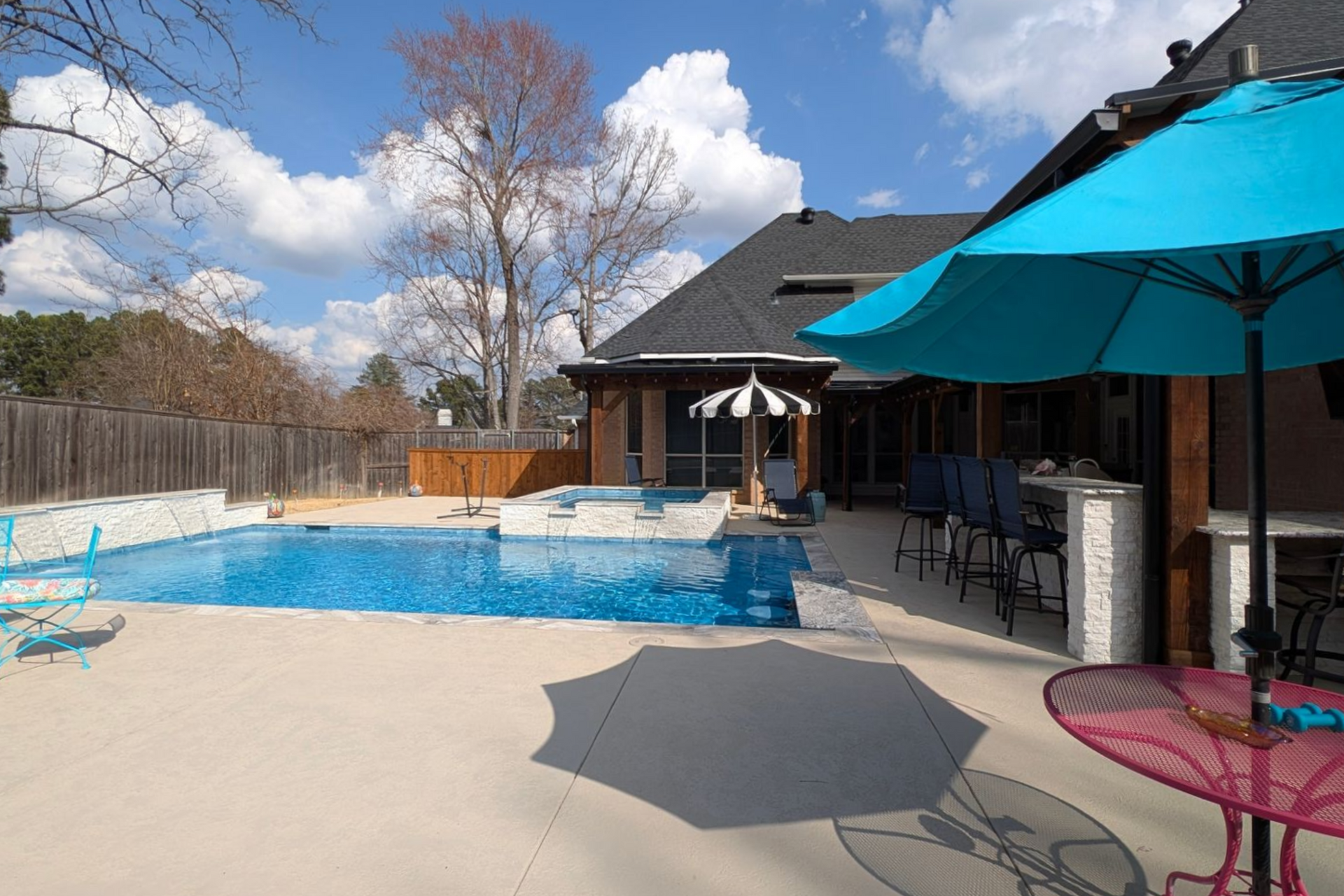 Backyard with a swimming pool, patio, and teal umbrella on a sunny day.