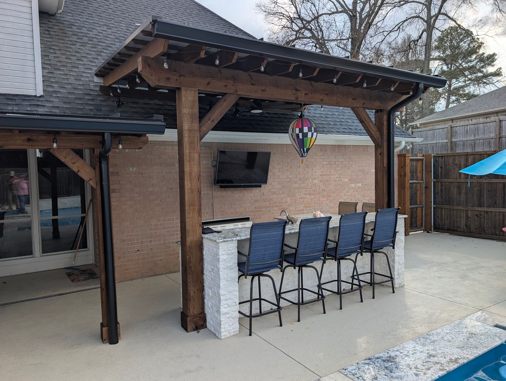Outdoor bar with seating, TV, and overhead structure. Brown wooden posts, white countertop, and black bar stools.