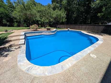 A water fountain bubbling in a backyard swimming pool with a stone wall and trees in the background.